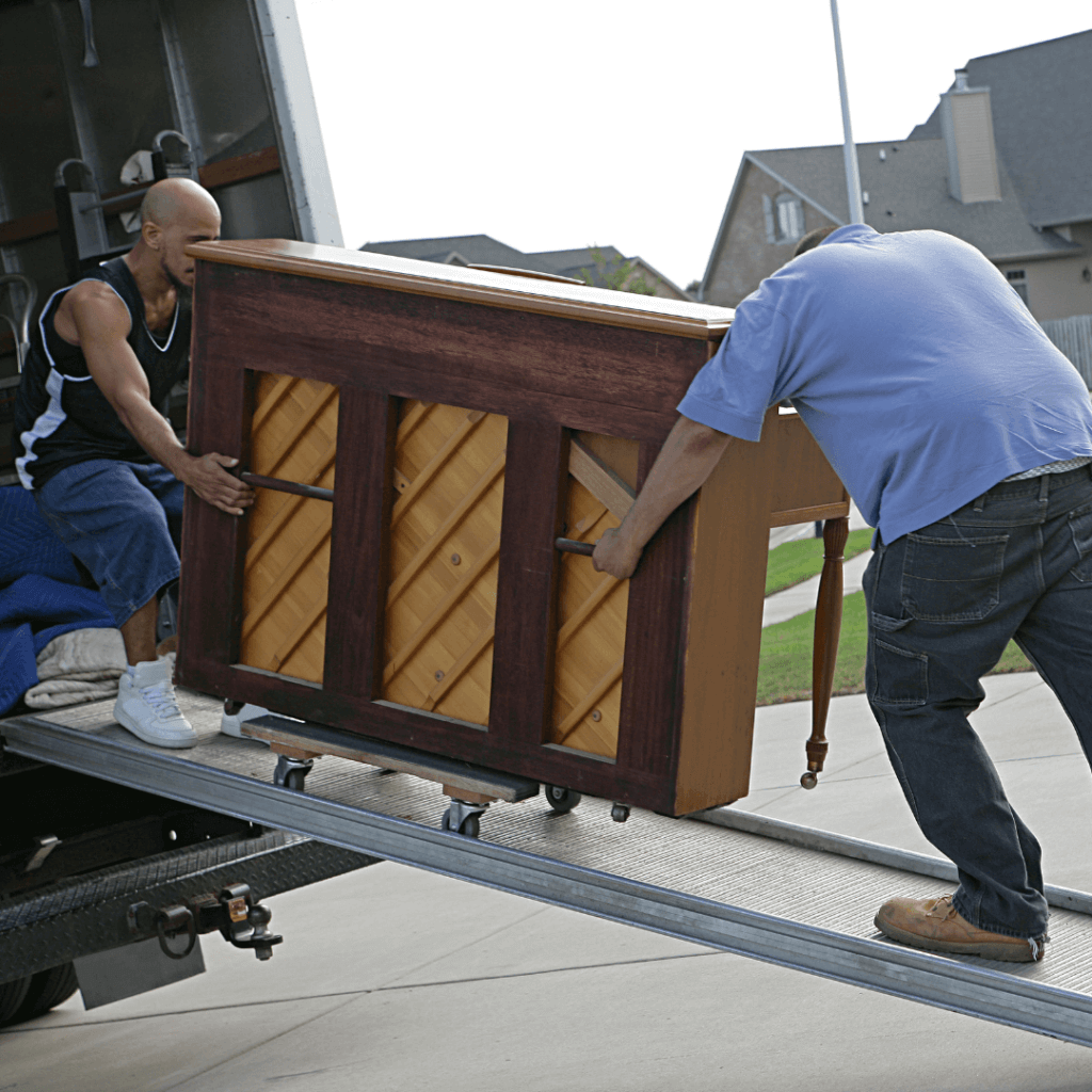 Two men rolling a brown upright piano down a ramp from a moving truck.